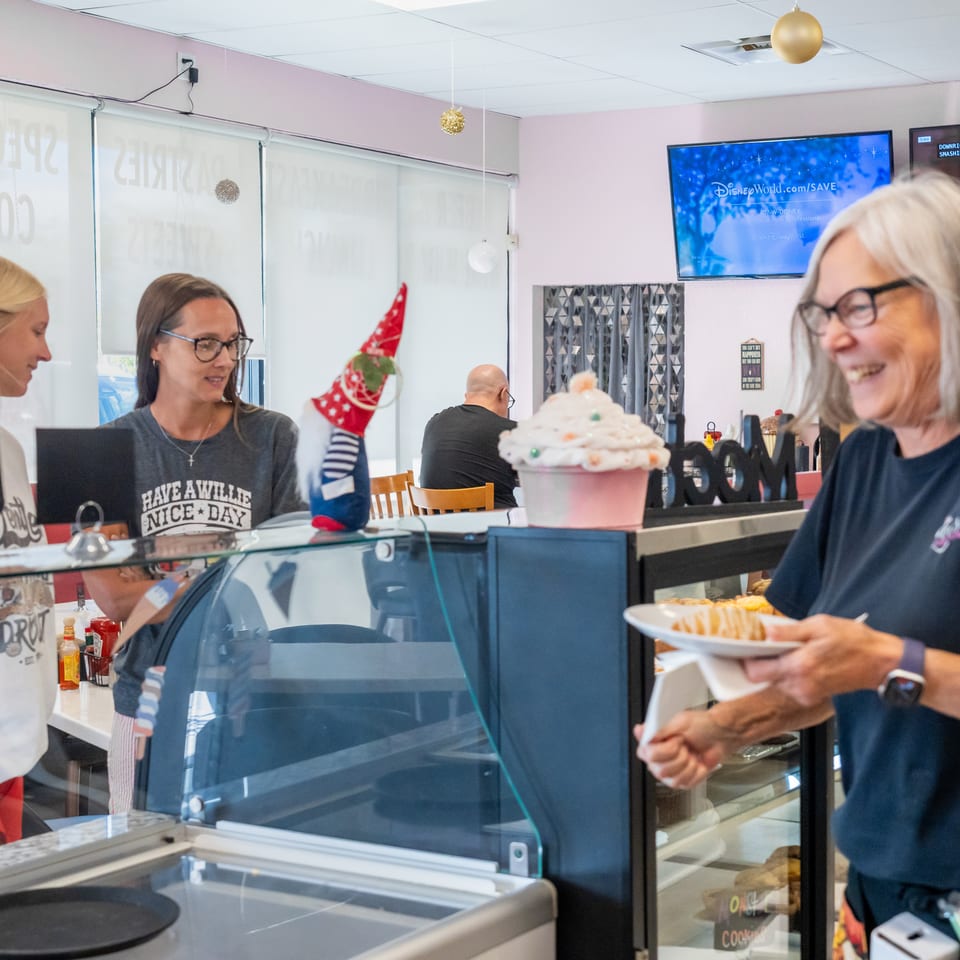 Restaurant staff serve a smiling customer holding a plate of fries beside a dessert display and ice cream counter.