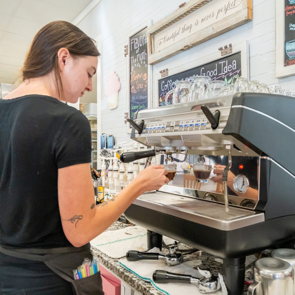 Barista prepares espresso at a commercial coffee machine beneath chalkboard menus in a bright café.