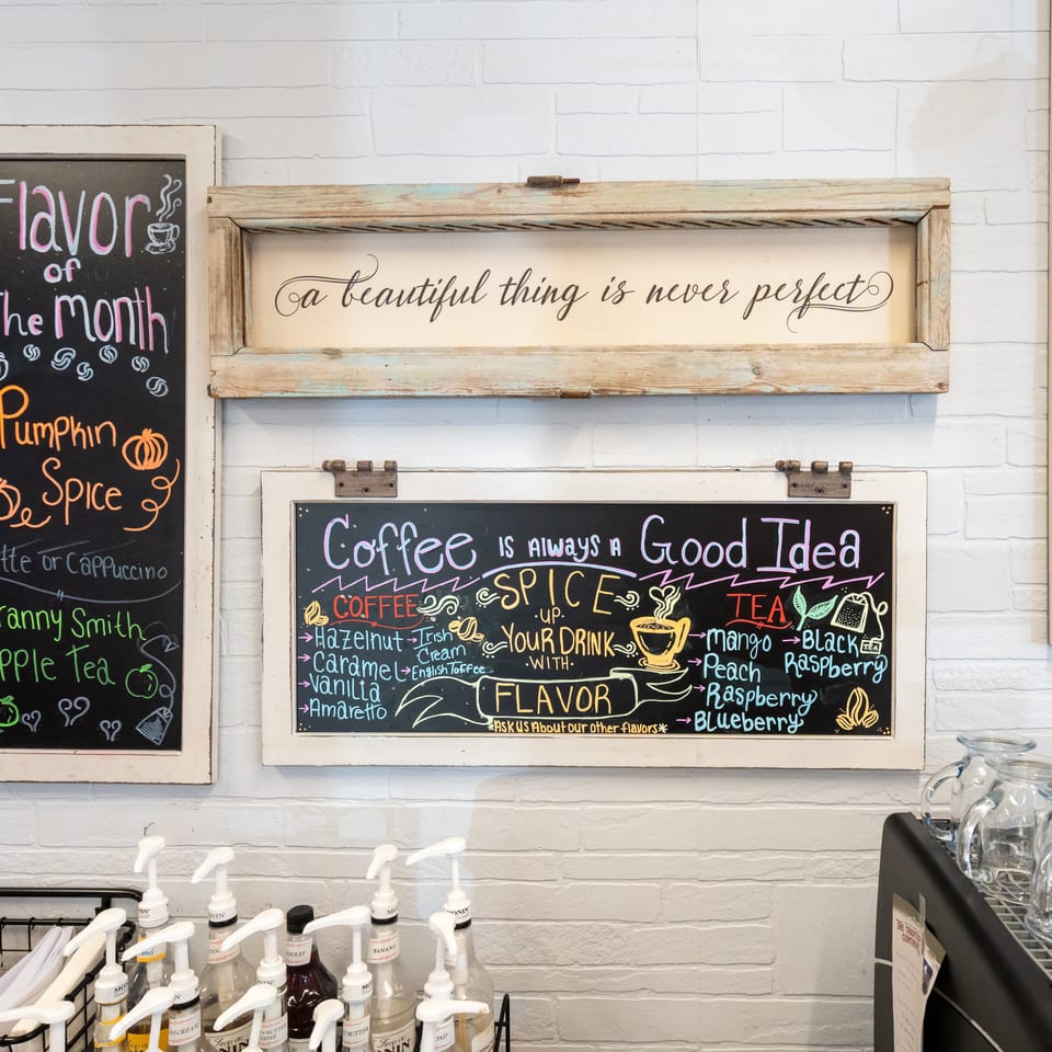 Coffee shop wall displaying colorful chalkboards about drink flavors, pumpkin spice, and coffee, with syrup pumps and glass pitchers below.