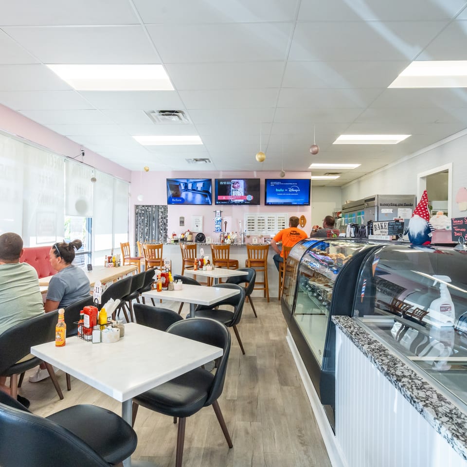 Bright café interior with white tables, black chairs, seated customers, televisions, dessert display case, and colorful chalkboard menus.