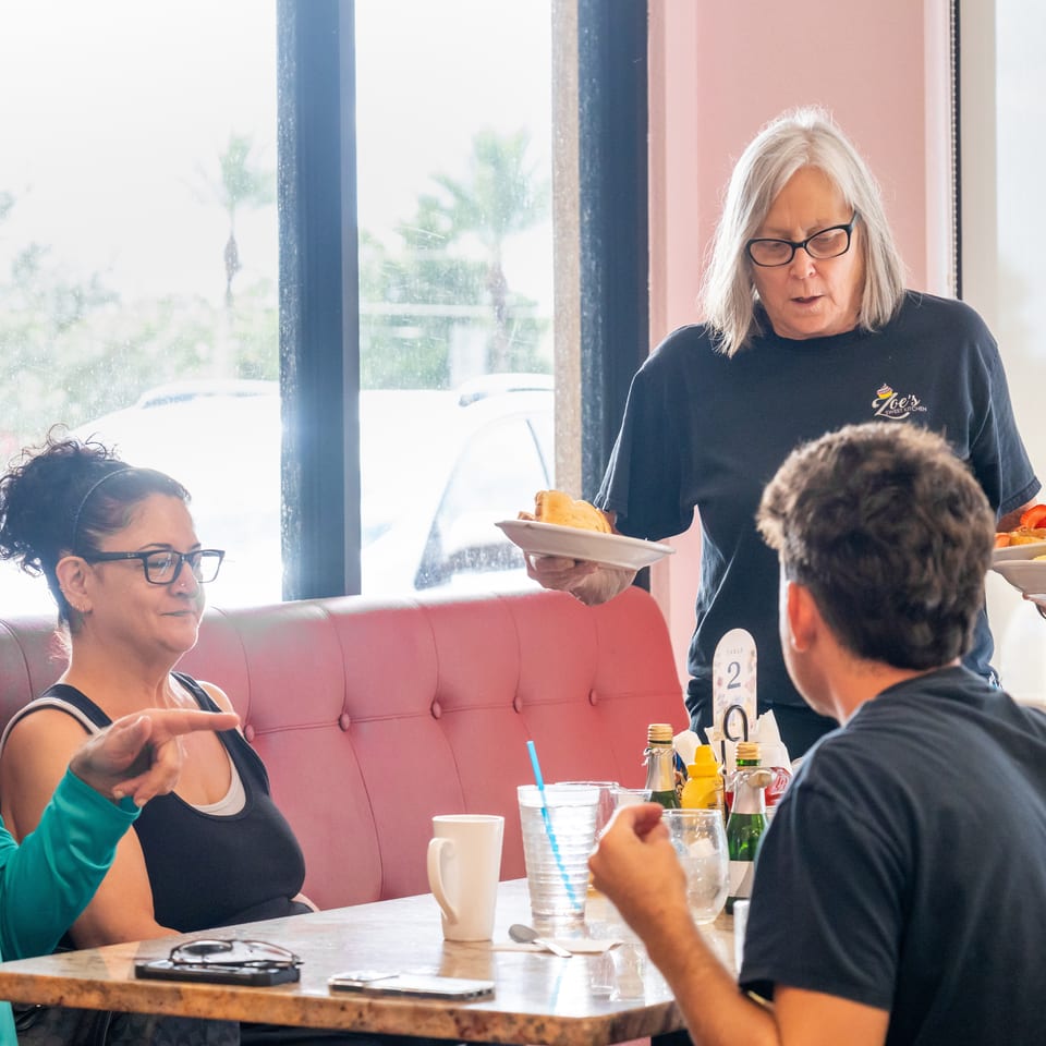Server carries plates of food to diners seated in a bright restaurant booth with pink upholstery.